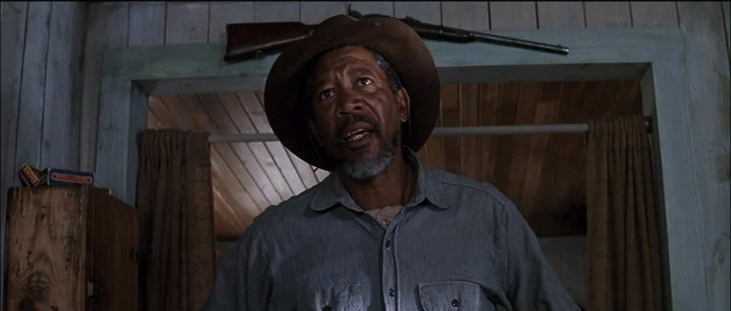 Unforgiven (1992) movie still: medium — A dimly lit room with wood-paneled walls and a rifle mounted above a doorway. A man in a hat stares forward…