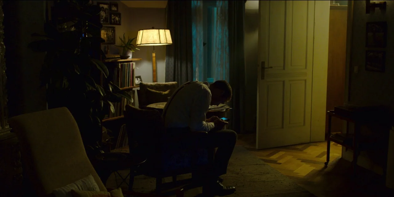 Mute (2018) movie still: medium — A dimly lit room with herringbone wood floors and a floor lamp emitting warm light. A man sits in a velvet…