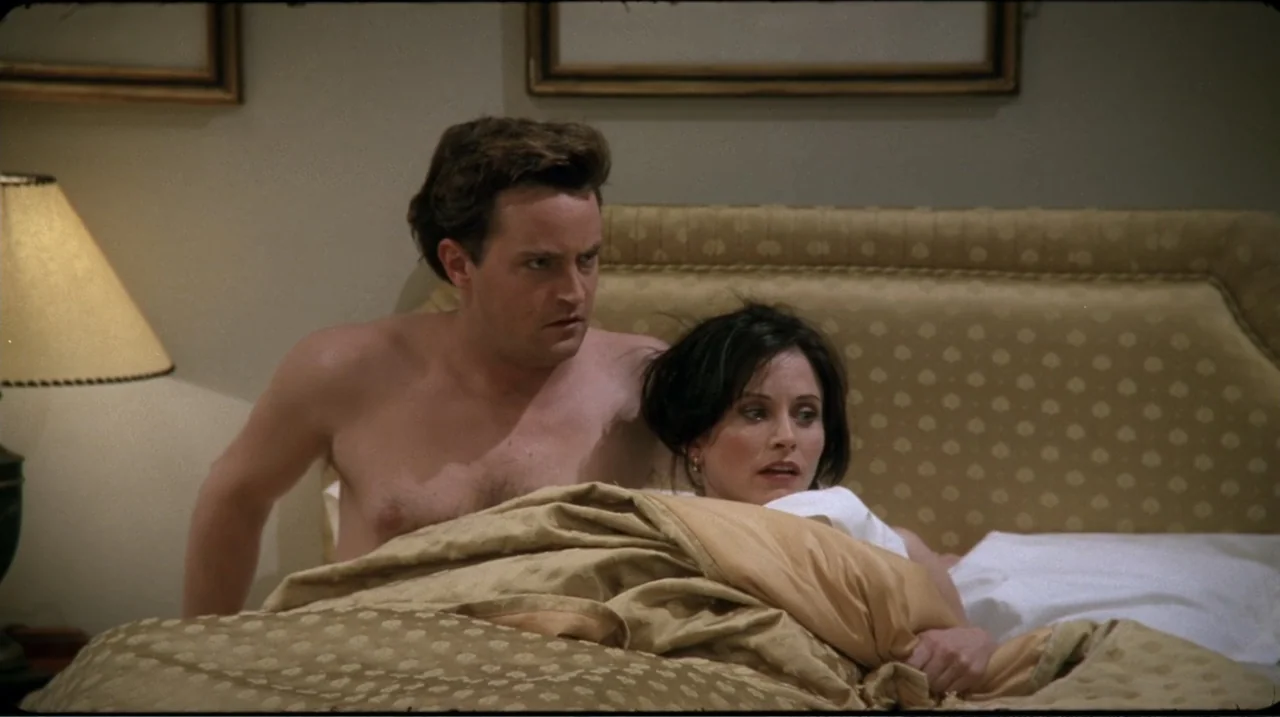 Friends: The Reunion (2021) movie still: medium — A furnished bedroom with a large beige headboard and a bedside lamp. A shirtless man and a woman…