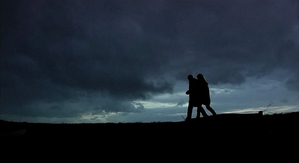 9 Songs (2004) movie still: wide — A vast, dark field under a heavy, overcast dusk sky. Two people in coats walk side by side across the flat…