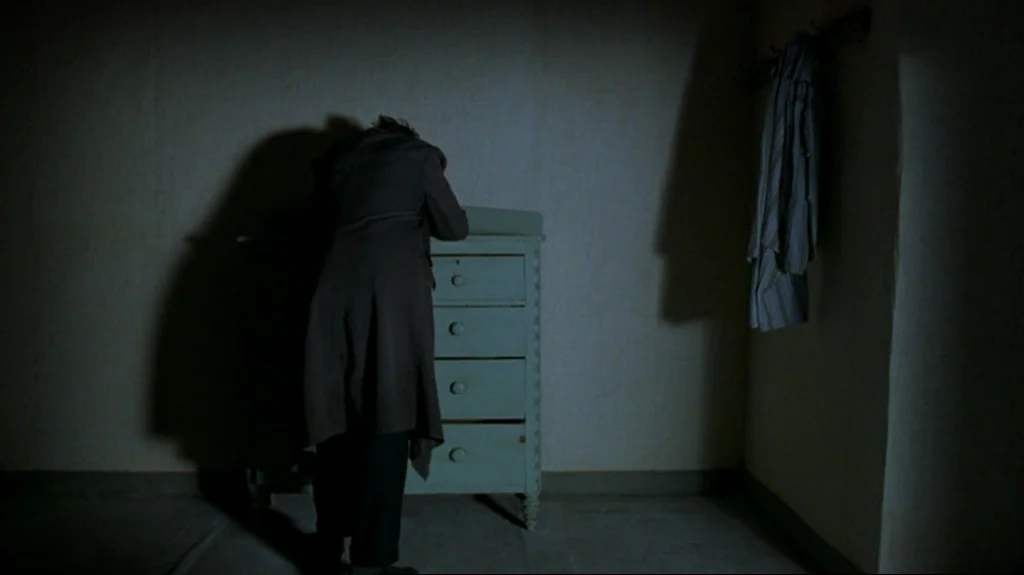Spider (2002) movie still: fullbody — A dimly lit room features a pale painted wooden chest of drawers. A lone figure stands facing the dresser…