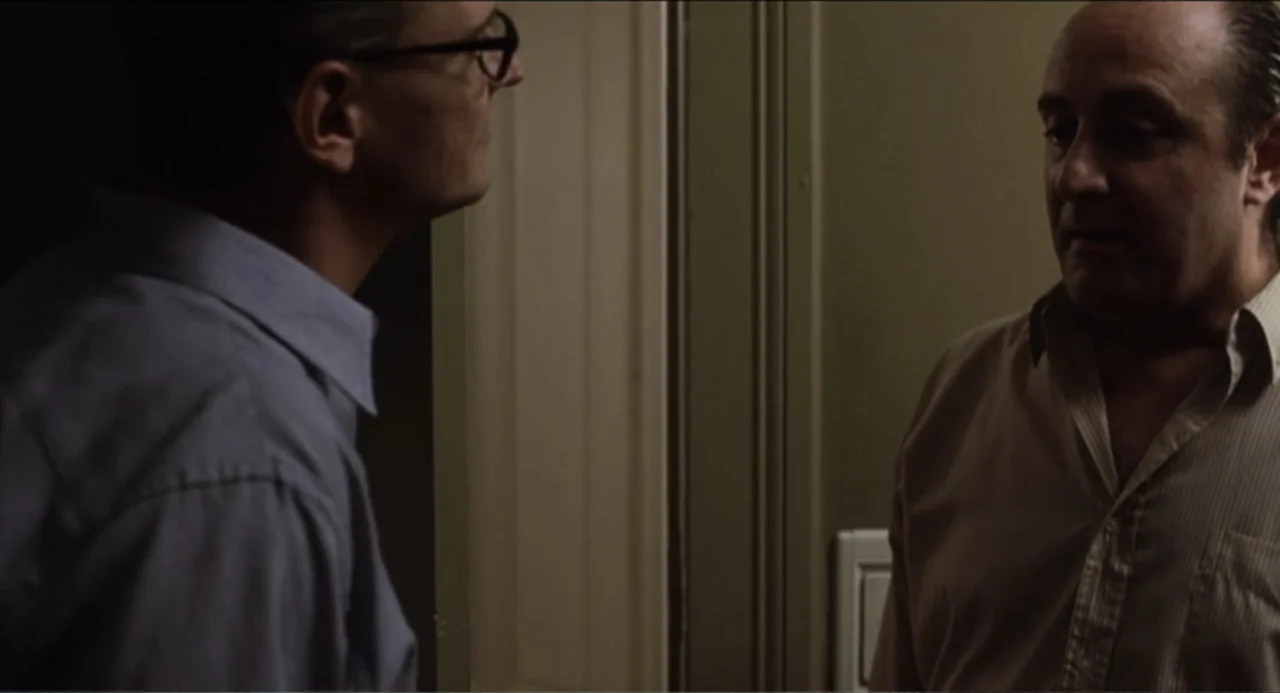 The Holy Girl (2004) movie still: medium — A dimly lit indoor space with a plain wall and a door frame. Two men in dress shirts stand near each…
