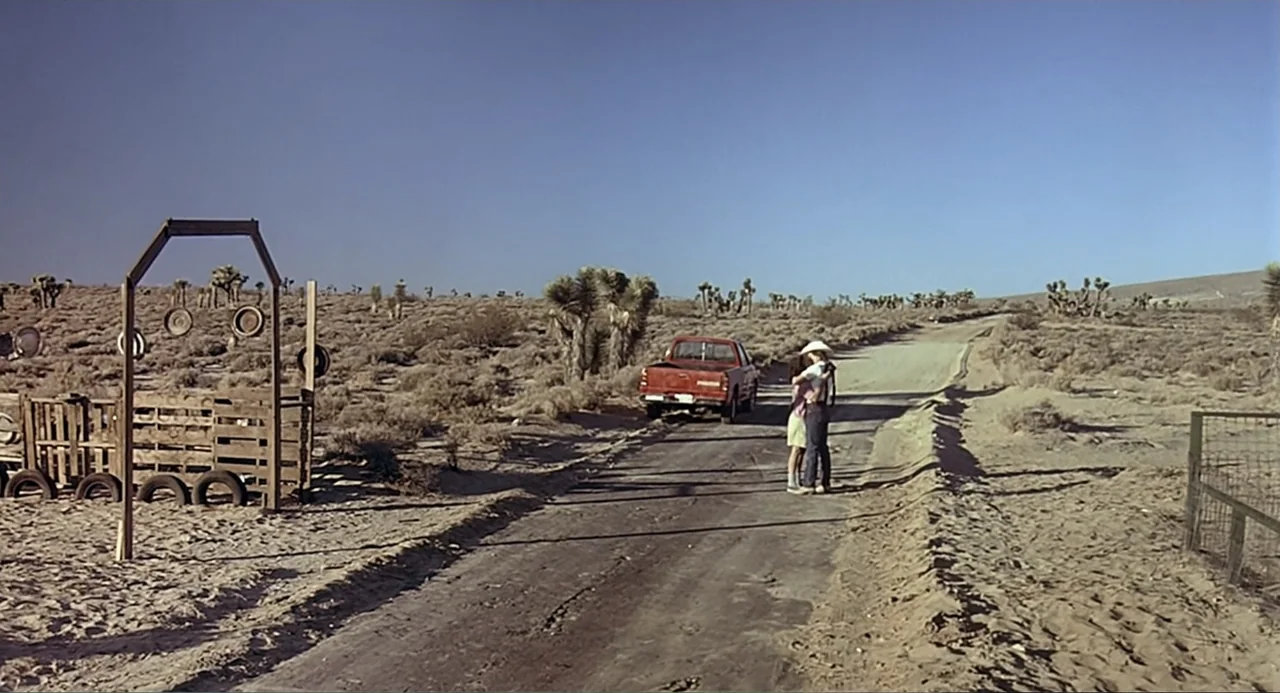 Tremors (1990) movie still: wide — A desolate desert road stretches through dry brush with a red pickup parked nearby. Two individuals stand on the…