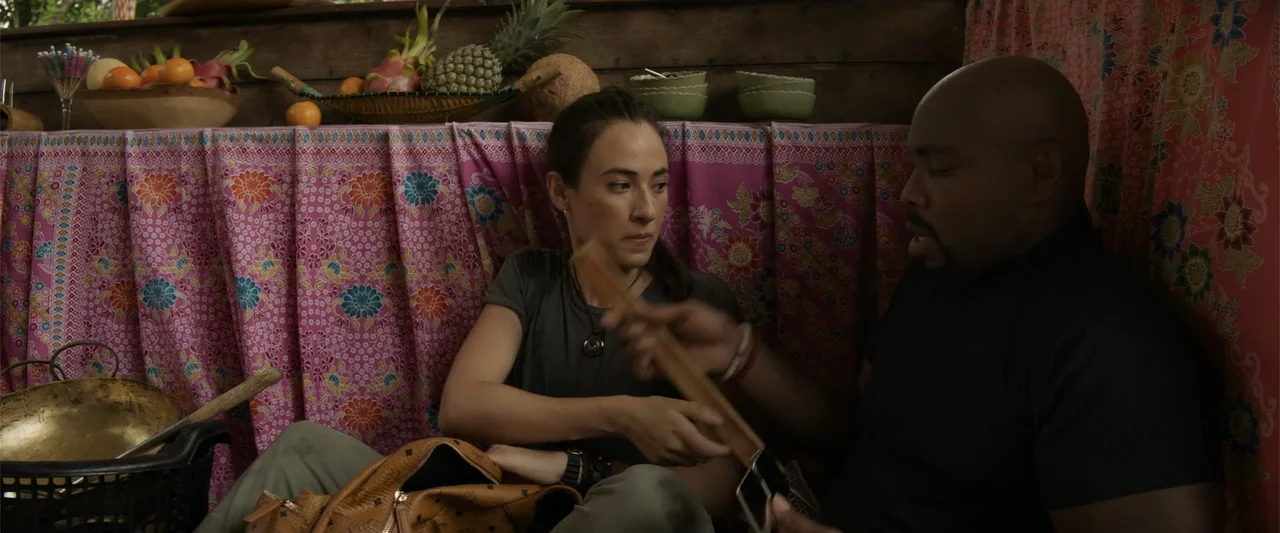 Meg 2: The Trench (2023) movie still: medium — A cramped stall features walls draped in vibrant pink patterned fabric and fruit displays. A woman…