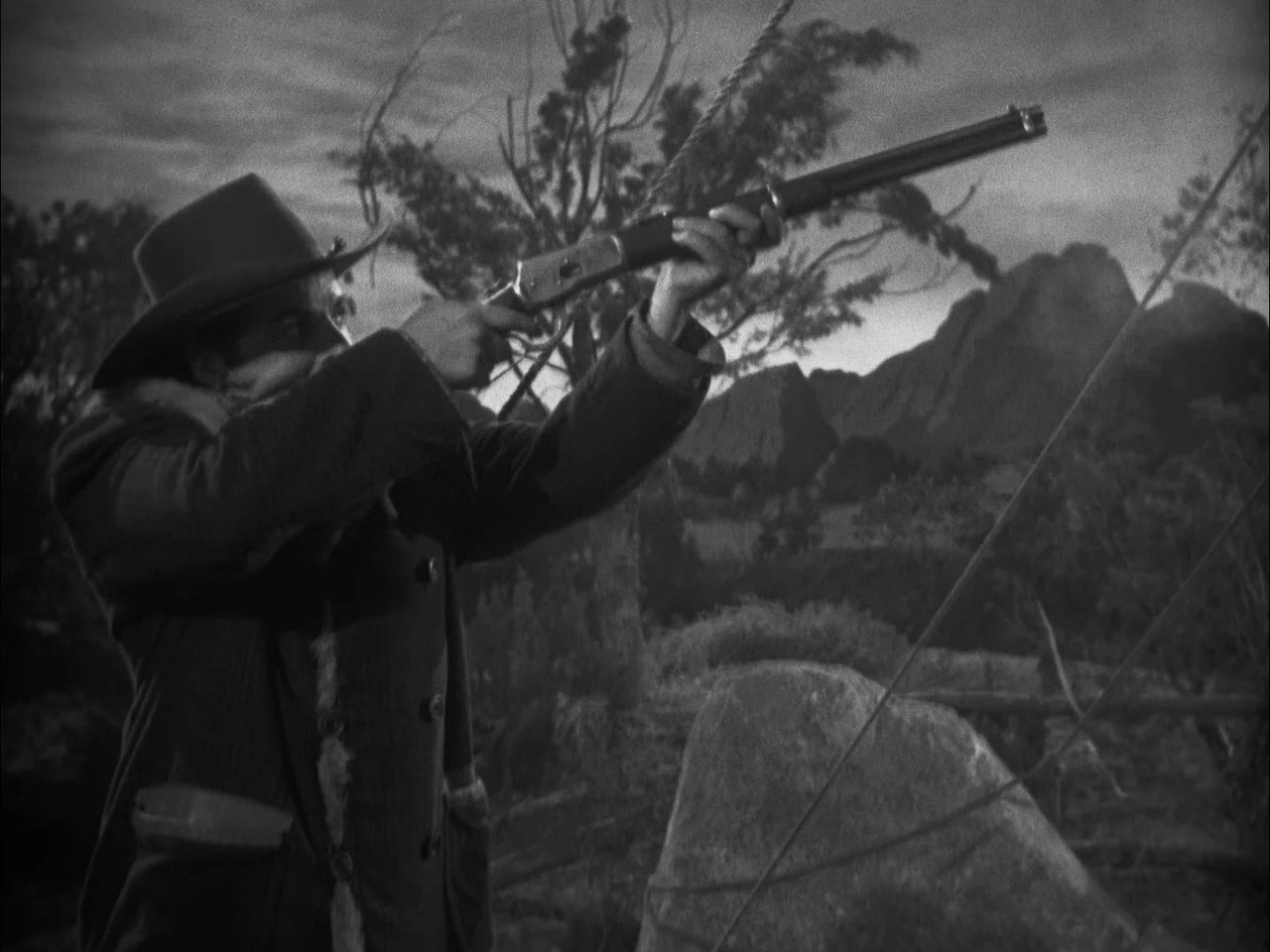The Ox-Bow Incident (1942) movie still: medium — A man in a brimmed hat and fur-lined coat stands in a rocky landscape. He holds a rifle and aims it…
