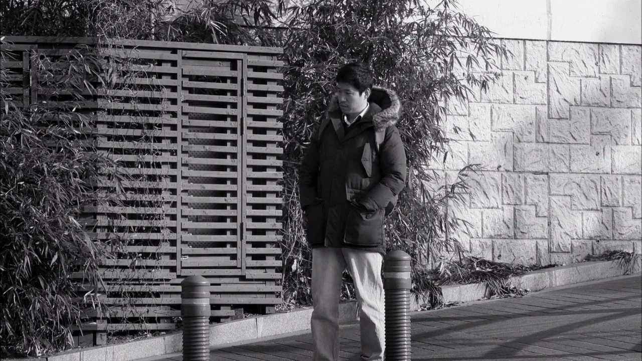 The Day He Arrives (2018) movie still: fullbody — A man stands on a paved sidewalk next to a wooden slatted structure. A light-colored stone…