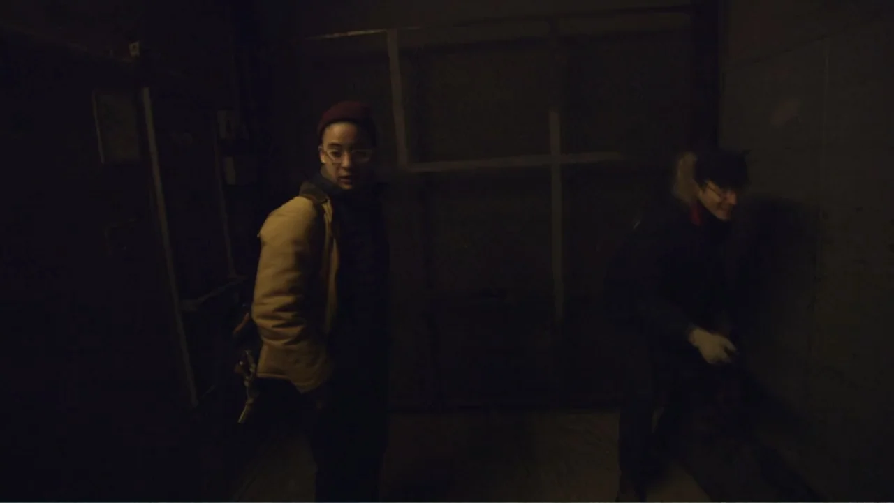 Suited (2016) movie still: medium — A dark, cramped interior with industrial features and concrete walls. Two people stand close together in the…