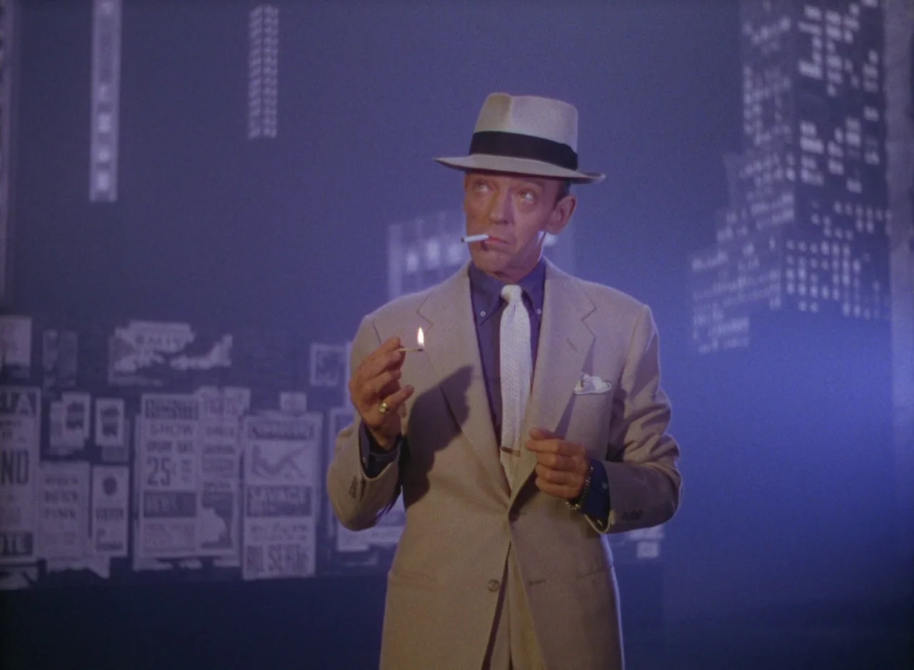 The Band Wagon (1953) movie still: medium — A man stands on a dark stage against a backdrop of blurry city posters. He holds a burning match to…