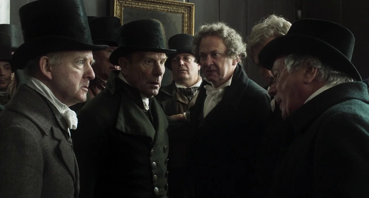 Peterloo (2018) movie still: medium — A group of Victorian men in dark formal attire gather in a dimly lit room. They stand close together while…
