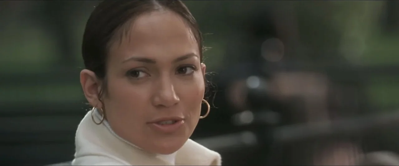 Maid in Manhattan (2002) movie still: closeup — An outdoor park setting with blurred greenery in the background behind a bench. A woman wearing a…