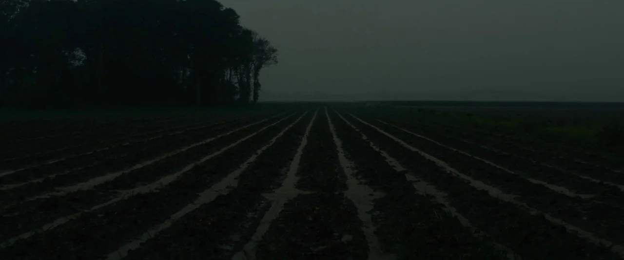 Mudbound (2017) movie still: establishing — A vast, plowed farm field stretches under a dark and overcast sky at night. Parallel rows of wet soil…