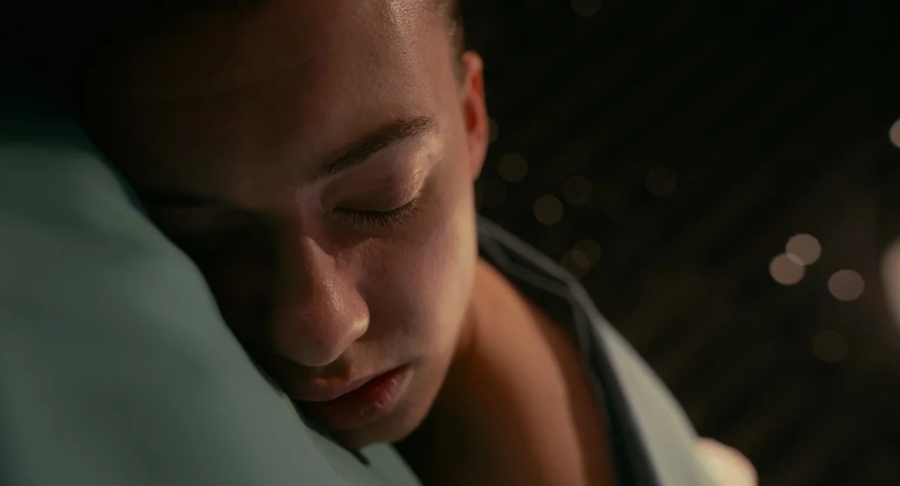 Fresh (2022) movie still: closeup — A dimly lit room provides a dark background with a few out-of-focus light spots. A person with closed eyes rests…
