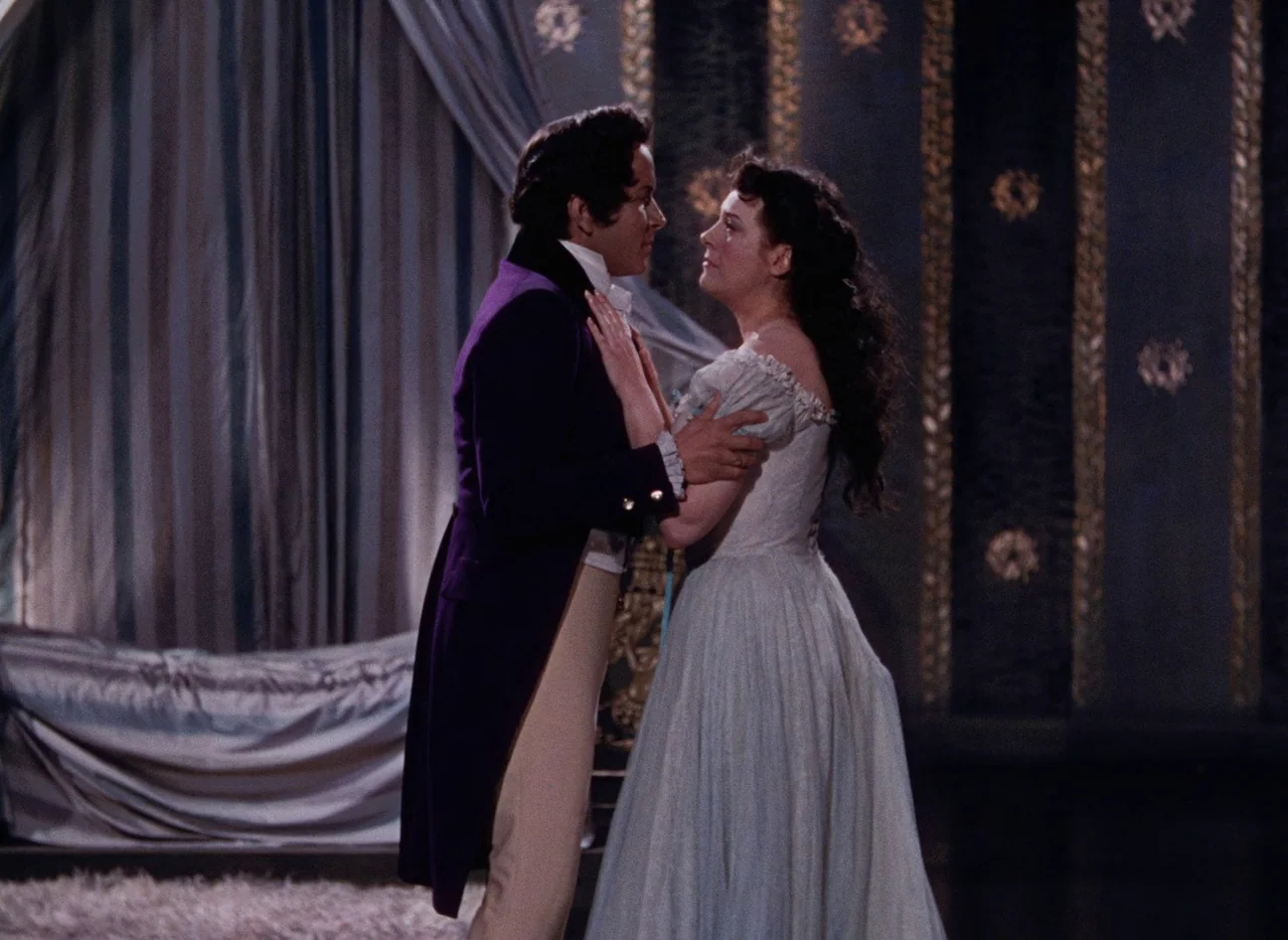 The Tales of Hoffmann (1951) movie still: fullbody — An opulent 19th-century bedchamber features dark patterned walls and heavy draped fabric…