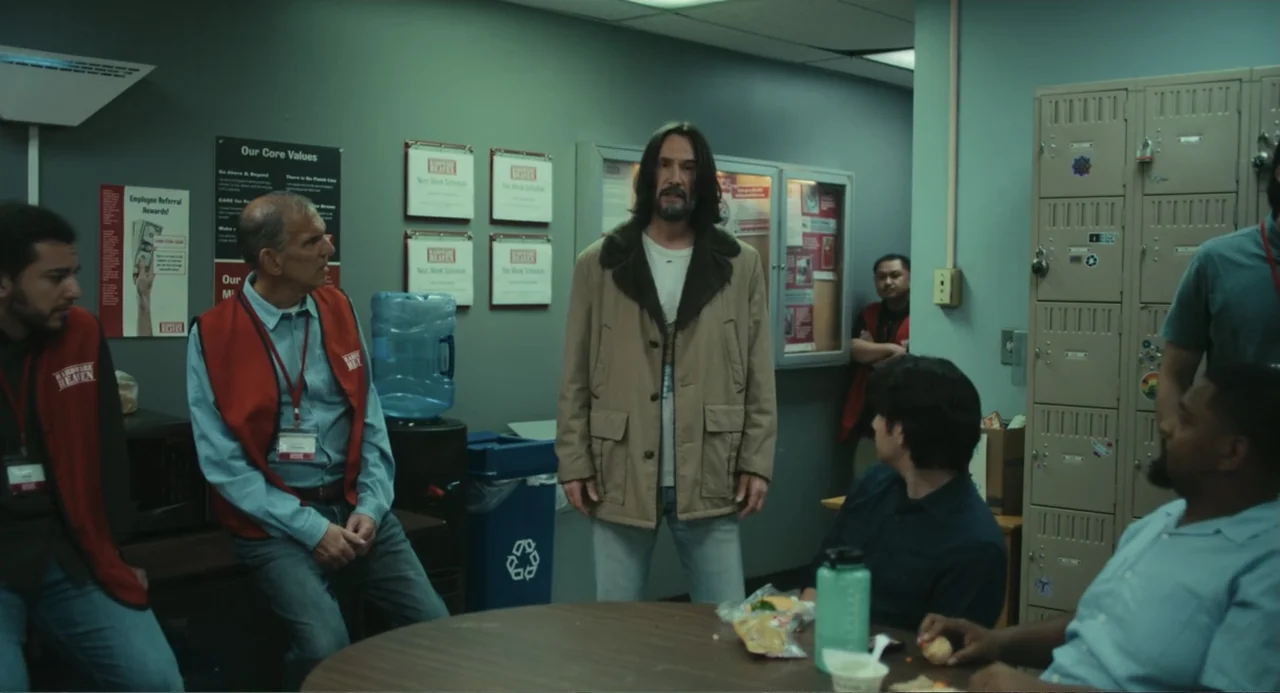 Good Fortune (2025) movie still: fullbody — A workplace breakroom features lockers, a water cooler, and posters on the green walls. Several…