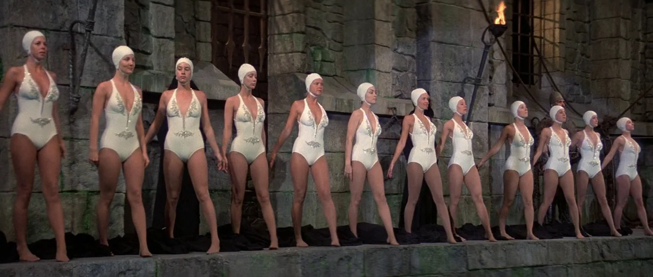 History of the World: Part 1 (1981) movie still: wide — A line of women in matching white swimsuits stands on a stone ledge. The background features…