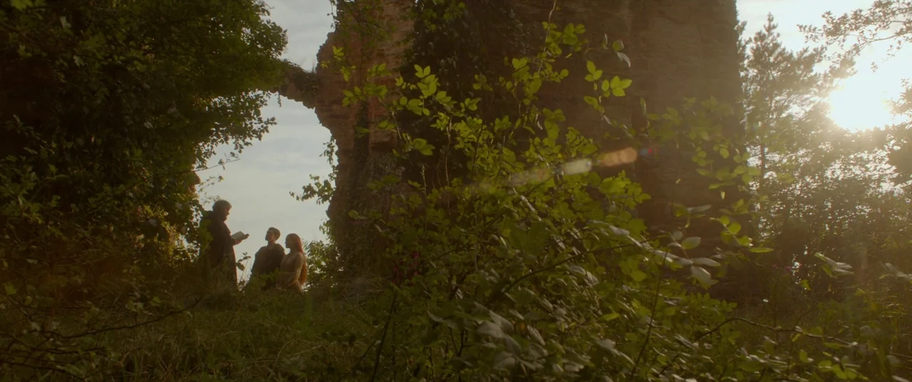 Ophelia (2018) movie still: wide — An overgrown stone archway stands amidst tall grass and trees in a forest. Three figures in period clothing stand…