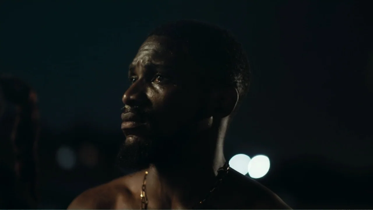Gangs of Lagos (2023) movie still: closeup — A shirtless man stands outdoors at night with blurry street lights in the distance. He gazes off-camera…