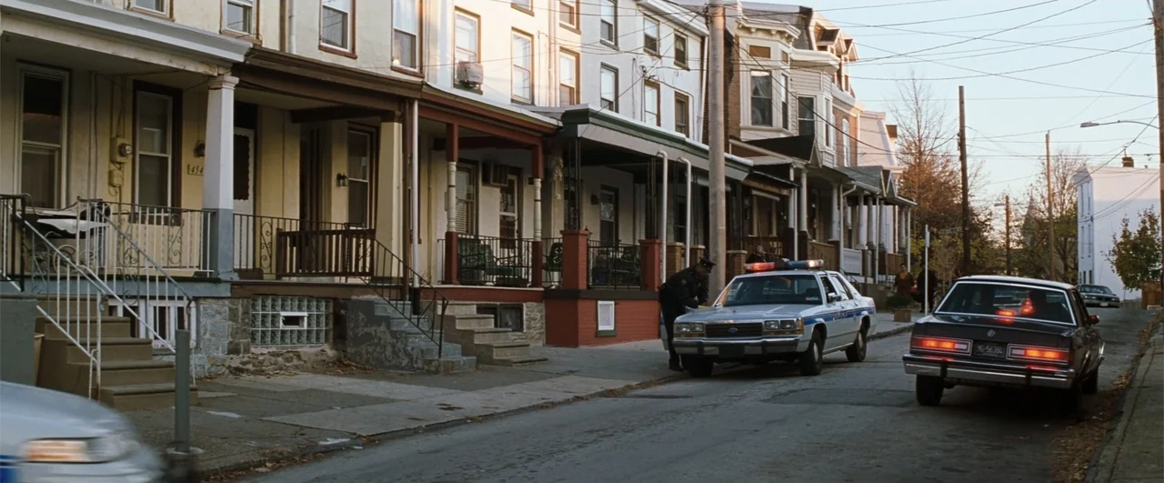 Fallen (2004) movie still: wide — A residential block of row houses features a street with a parked police cruiser. An officer in uniform leans…