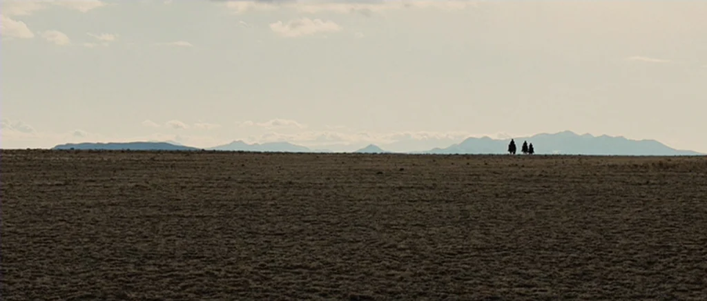 True Grit (2010) movie still: wide — A flat, expansive desert floor stretches toward a distant, low mountain range under clouds. Three small figures…