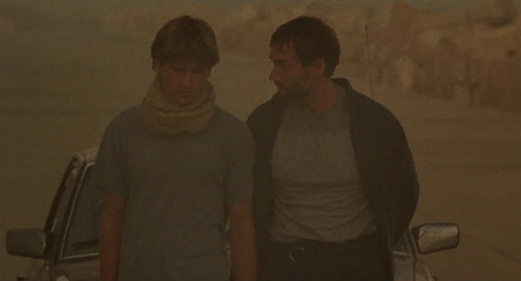 Furia (1999) movie still: medium — An arid, dust-filled landscape obscured by a thick, yellowish atmospheric haze. Two men stand close together in…