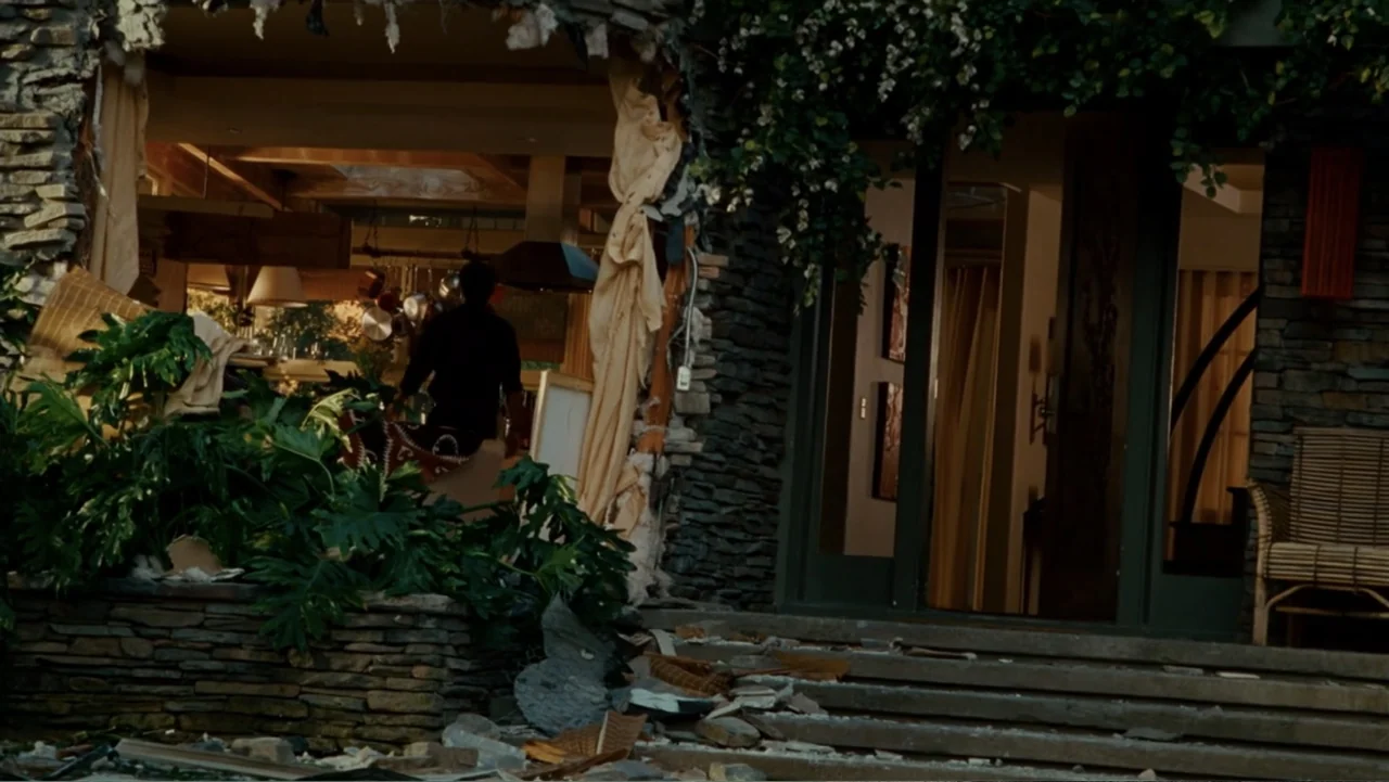 Hancock (2008) movie still: wide — A stone-walled residential exterior features a massive structural breach revealing an interior room. A man stands…