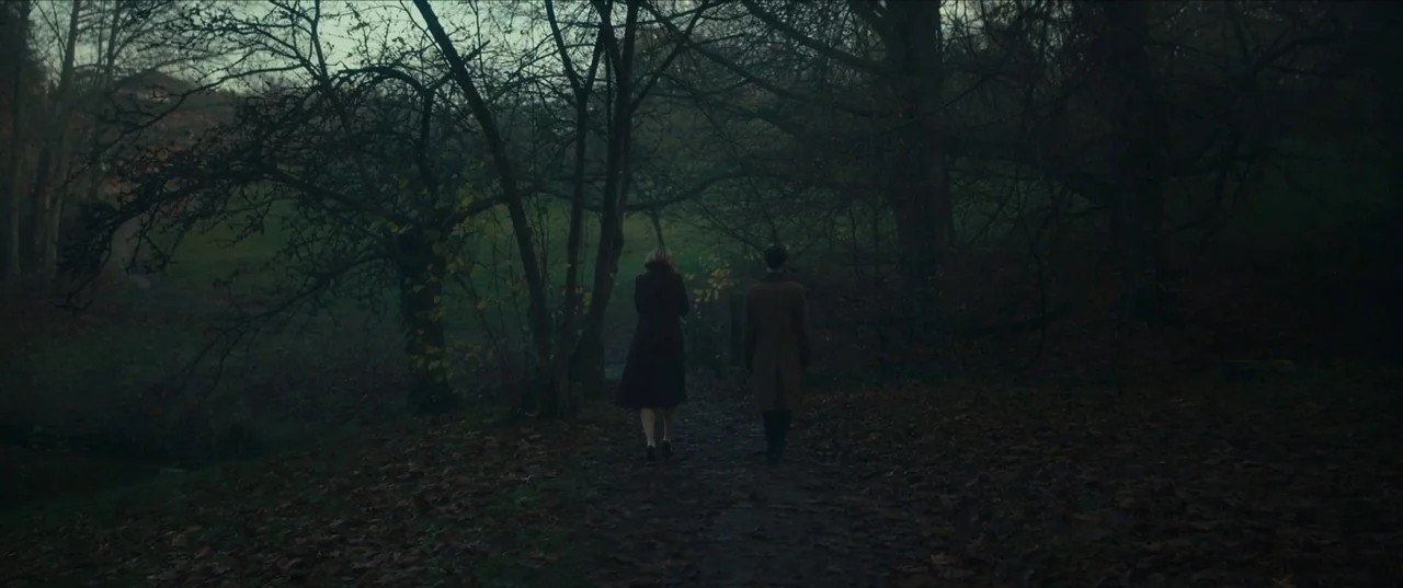 Earwig (2022) movie still: wide — A damp dirt path winds through a dense, dimly lit forest of dark trees. A man and a woman in mid-century coats…