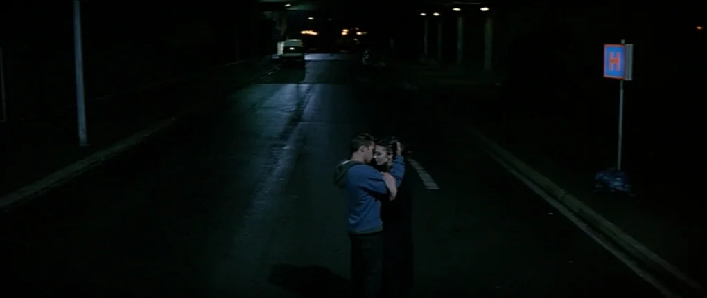 IP5 (1992) movie still: wide — A dark, empty asphalt road stretches under a bridge at night. A young couple stands in the center of the road…