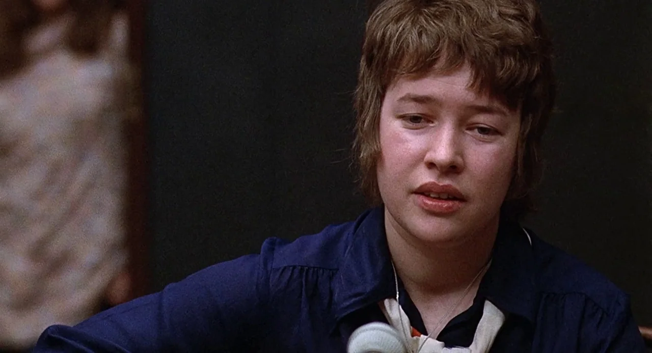 Taking Off (1971) movie still: medium — A person stands in a dimly lit, dark-walled courtroom setting. They speak clearly into a silver microphone…
