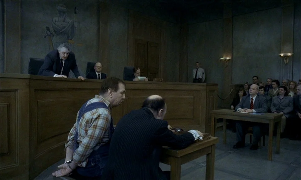 You, The Living (2007) movie still: wide — A dimly lit room with wood-paneled walls and a painted mural of Lady Justice. A man in handcuffs sits at…