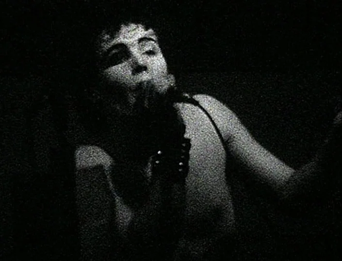 Glitterbug (1994) movie still: medium — A dark, minimally lit space with heavy grain texture. A performer sings into a microphone while facing…
