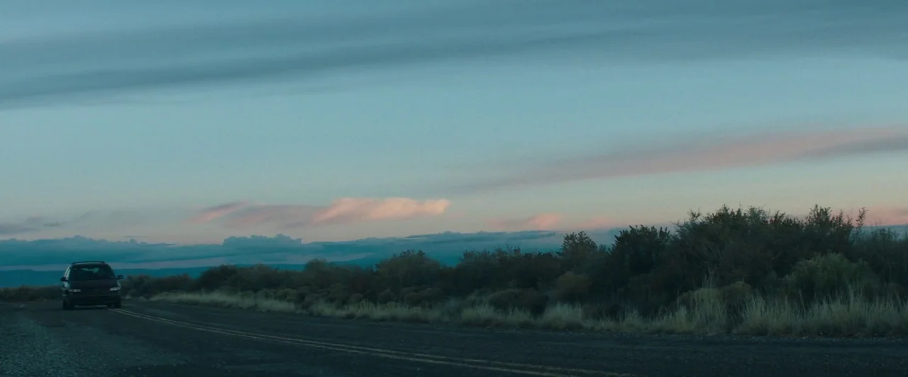 The Long Dumb Road (2018) movie still: wide — A long, flat rural road cuts through a dry desert scrubland at twilight. A single dark minivan travels…