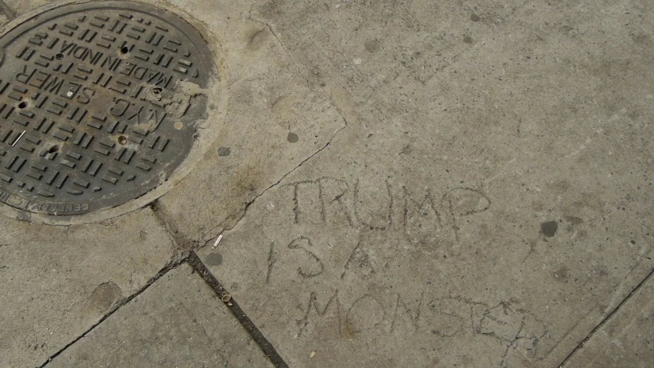 Counting (2015) movie still: wide — A weathered gray concrete sidewalk features a circular cast iron sewer grate. The words TRUMP IS A MONSTER are…