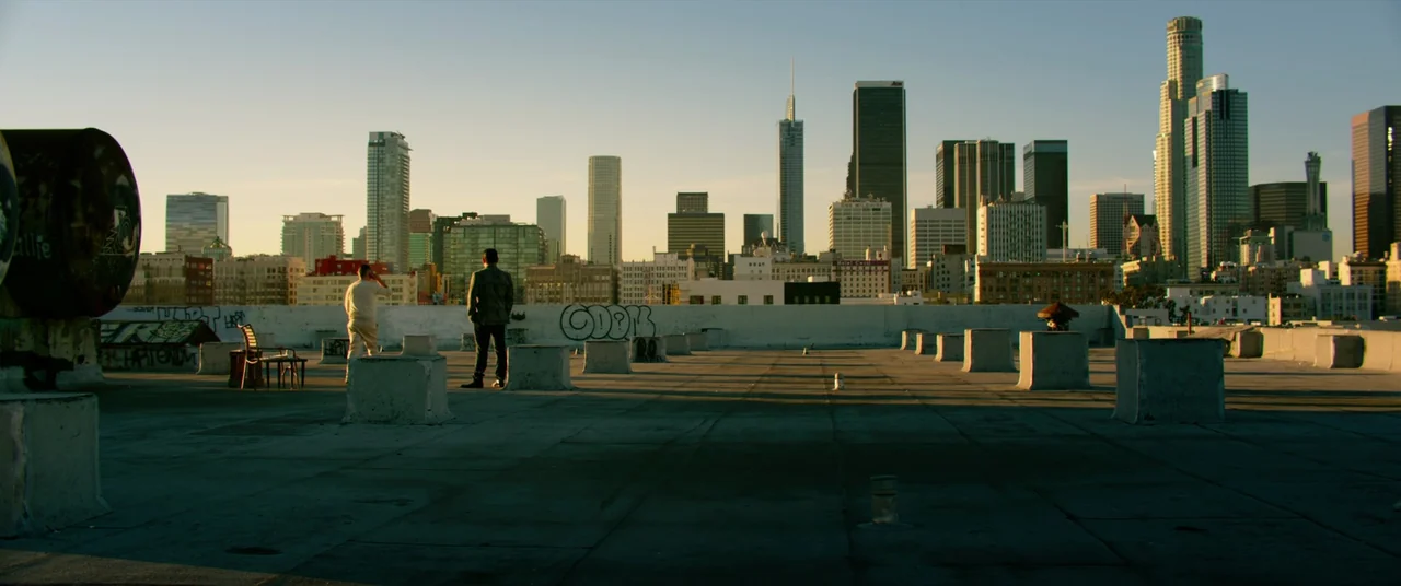 Ambulance (2022) movie still: wide — A flat concrete industrial rooftop features graffiti on a low perimeter wall at sunset. Two men stand looking…