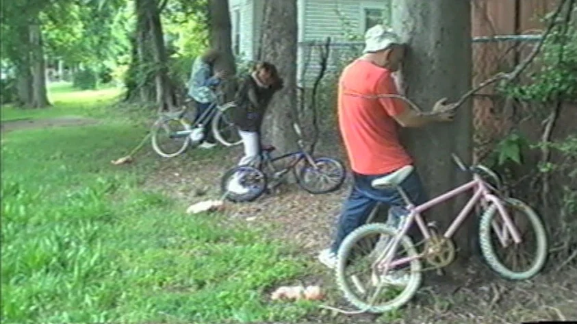 Trash Humpers (2009) movie still: wide — A grassy yard with several trees and a chain-link fence borders a house exterior. Three individuals lean…