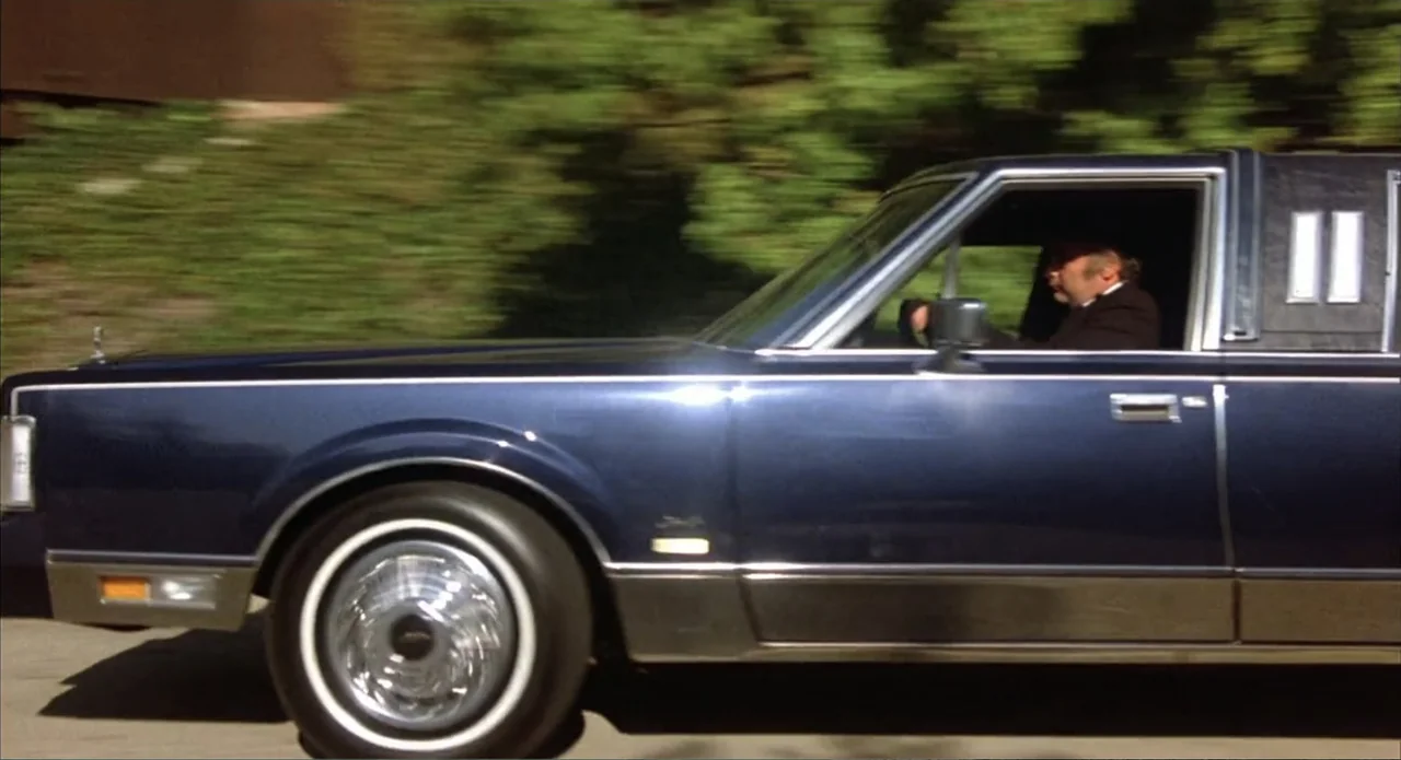 Back to School (1986) movie still: medium — A dark blue Lincoln Town Car travels along a street with blurred green foliage nearby. A man in a suit…