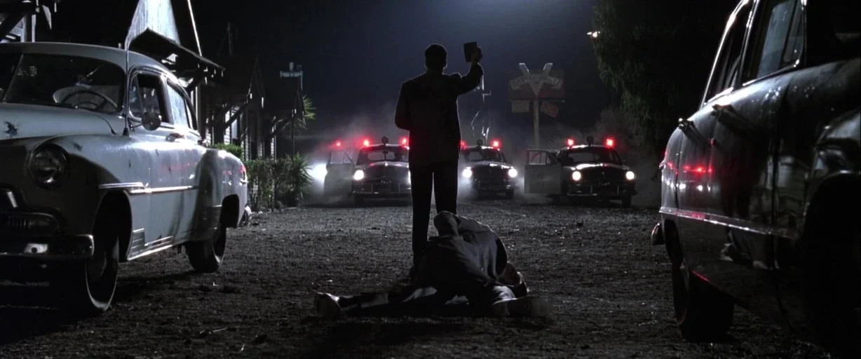 L.A. Confidential (1997) movie still: wide — A dark, unpaved lot is surrounded by vintage 1950s automobiles with illuminated police lights. A man in…