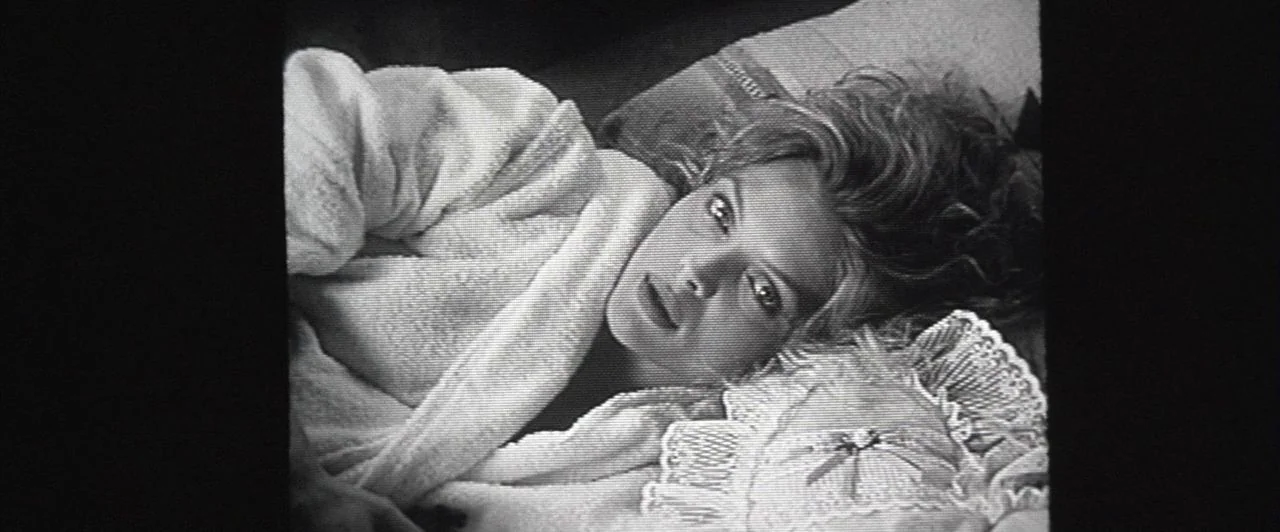 The Witches of Eastwick (1987) movie still: closeup — A woman rests on a bed covered with patterned linens and decorative pillows. She lies on her…