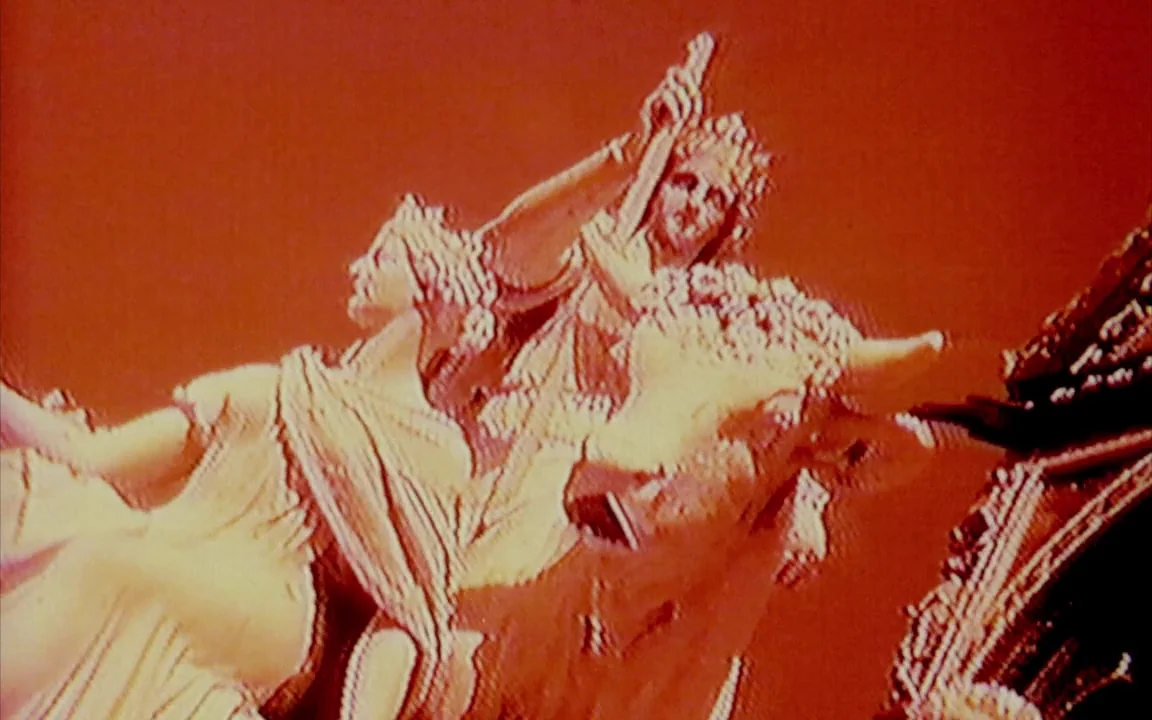 The Last of England (1987) movie still: medium — A stone sculpture of two classical figures sits against a dark red backdrop. The figures appear to…