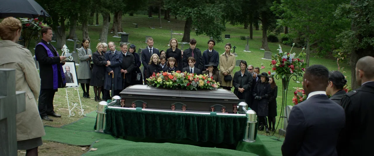 The Empty Man (2020) movie still: wide — A group of mourners stands around a casket on a green carpet in a cemetery. A priest leads the service…