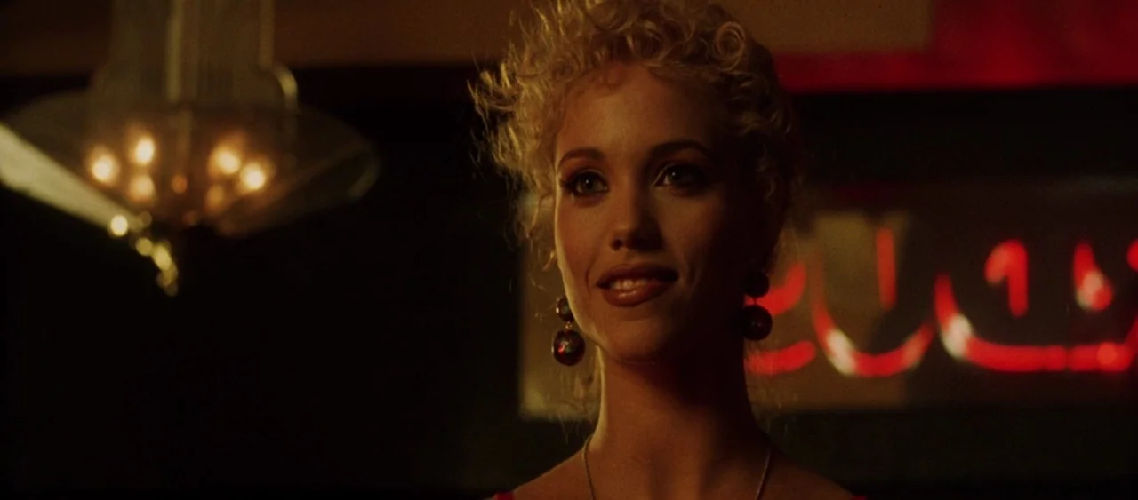 Showgirls (1995) movie still: closeup — A dimly lit bar features a red neon sign and an out-of-focus chandelier. A young woman with curly blonde…