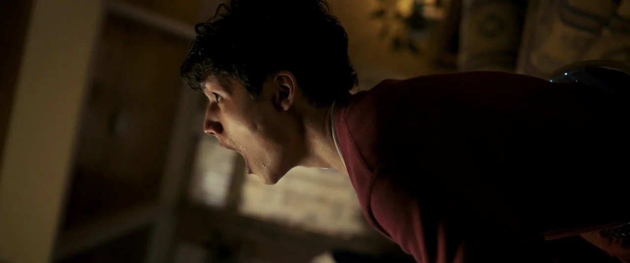 Talk To Me (2022) movie still: medium — A dimly lit room provides a dark background behind the subject. A young man stares directly ahead with his…