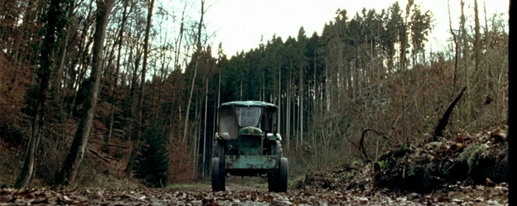 Calvaire (2004) movie still: wide — A rustic tractor sits centered on a dirt path between rows of dense trees. Dead leaves cover the ground as the…