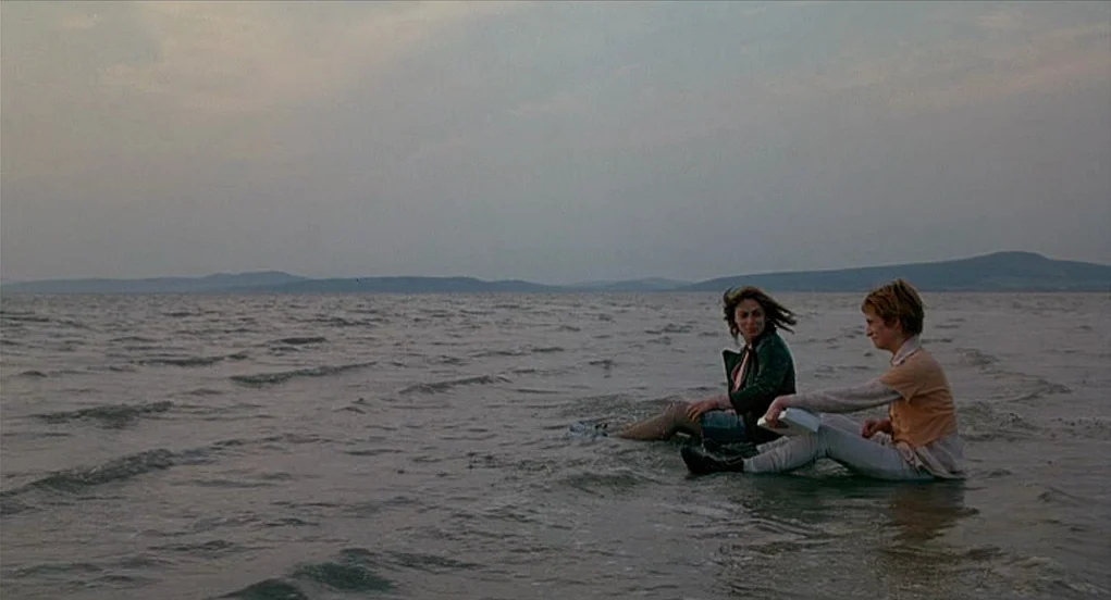 Butterfly Kiss (1995) movie still: wide — A vast, calm lake under a heavy, grey overcast sky with distant rolling hills. Two women sit partially…