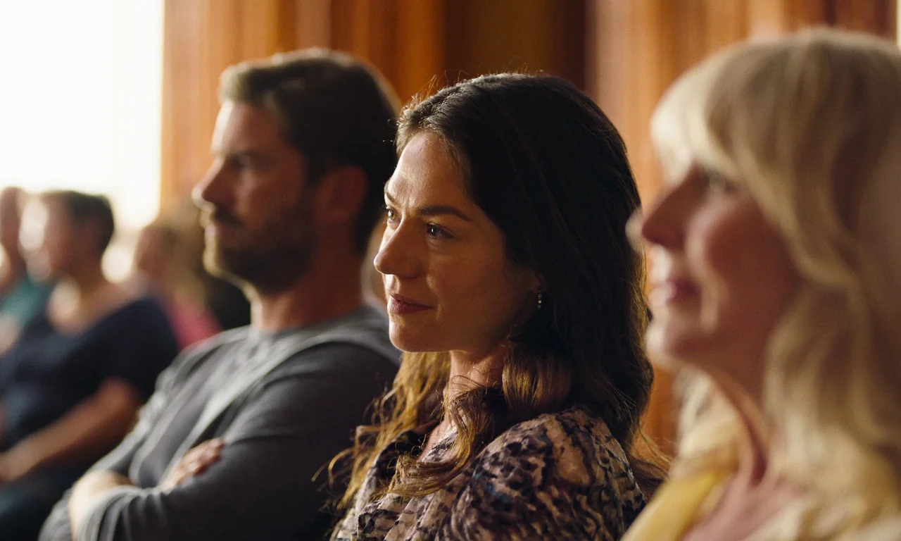 Close (2022) movie still: medium — An interior space with wooden paneled walls behind a seated audience. Several people sit in rows, focusing their…