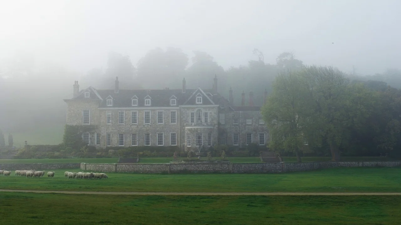 Emma. (2020) movie still: establishing — A stone country manor house sits behind a low wall in thick morning fog. A small herd of sheep grazes on…
