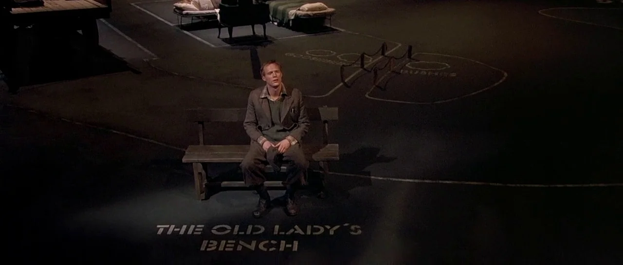 Dogville (2003) movie still: wide — A dark, minimalist theatrical stage features chalk lines on the floor and small cots. A man sits alone on a…