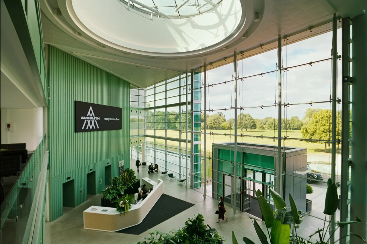 Bugonia (2025) movie still: establishing — A spacious, modern corporate atrium features mint-green ribbed walls and a circular skylight above. A…