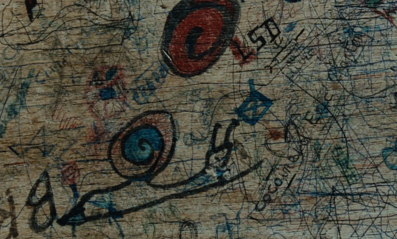 Graduate First (1978) movie still: closeup — A light brown wooden surface is densely covered with chaotic pen and marker graffiti. Scratched-in…