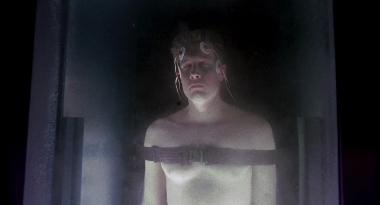 Night of the Creeps (1986) movie still: medium — A man stands inside a narrow vertical glass cylinder filled with faint vapor. He is held upright by…