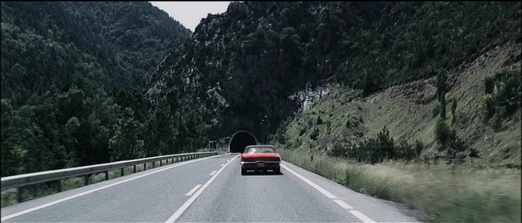 The Machinist (2004) movie still: wide — A rural asphalt highway winds between steep, forest-covered mountains towards a dark tunnel mouth. A single…